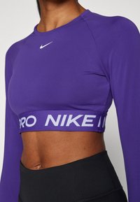 Lilla langermet treningscrop-top laget av elastisk stoff, med en hvit Nike-logo og et elastisk bånd med hvite detaljer og teksten "PRO".