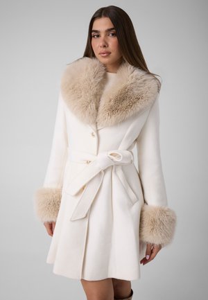 Femme portant un manteau blanc avec un large col en fourrure beige et des poignets en fourrure assortis, attaché à la taille avec une ceinture.