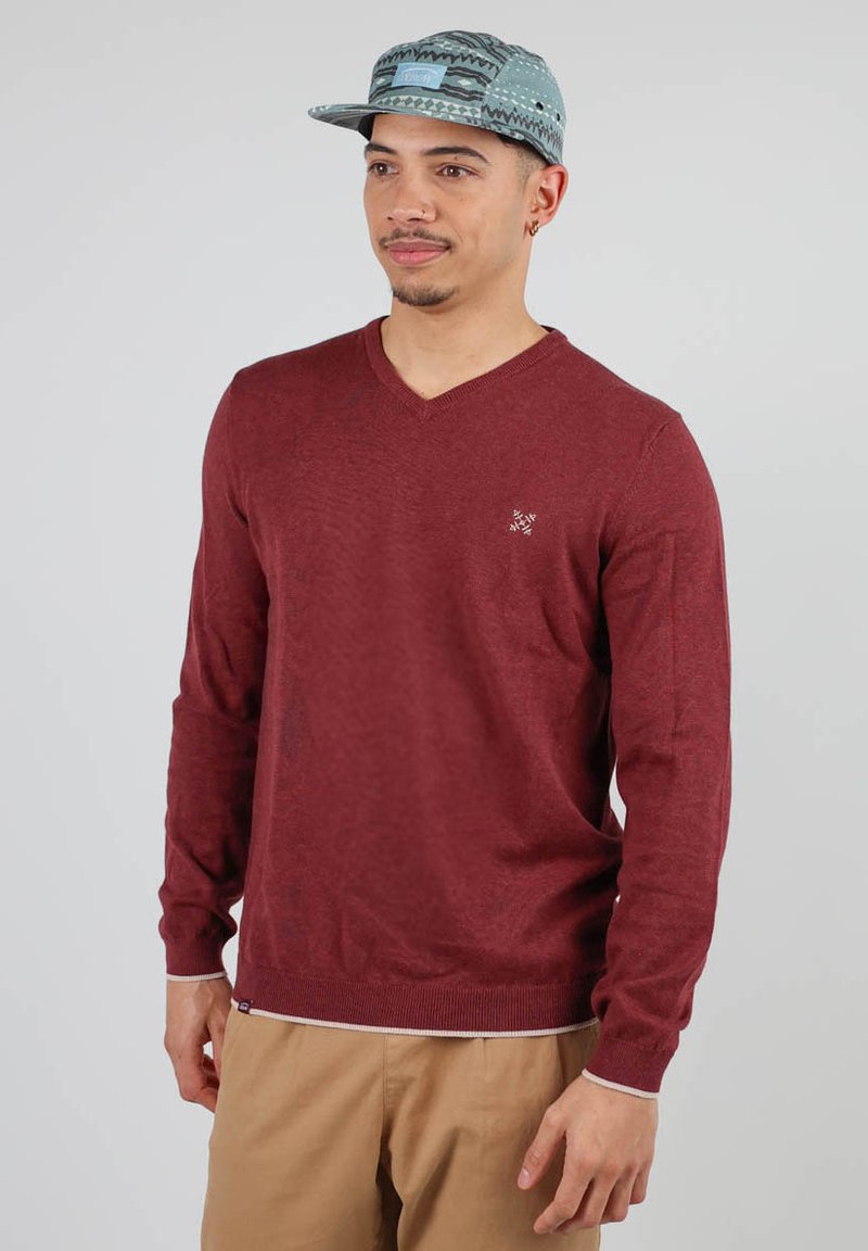 Pull à col en V bourgogne avec manches longues, en tissu tricoté texturé, logo brodé sur la poitrine, associé à un pantalon kaki et une casquette à motifs.