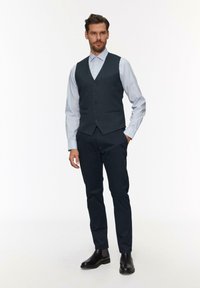 Marine vest over een lichtblauw shirt, slim fit, vijf knopen, zijzakken, gecombineerd met donkere broek en zwarte Chelsea boots.