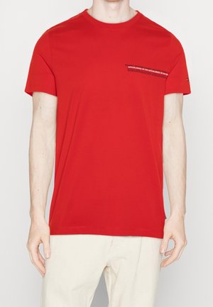 Camiseta de algodón roja con cuello redondo, mangas cortas y un bolsillo en el pecho que presenta un detalle de texto. Textura suave y corte regular.