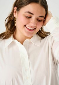 White button-up shirt with a classic collar, featuring a smooth texture and pearl-like buttons. Hair styled down, with gold earrings.