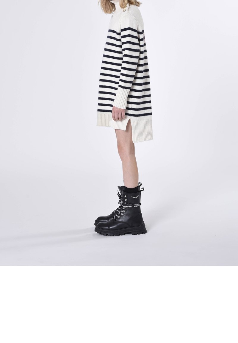 Personne portant une robe-pull longue blanche et noire à rayures avec une fente latérale et des bottes de combat noires à lacets, vue de profil.