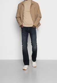 Béžová lehká bunda, béžový svetr, tmavé džíny z denimu a bílé tenisky s hnědými detaily. Model stojí s rukama v kapsách.