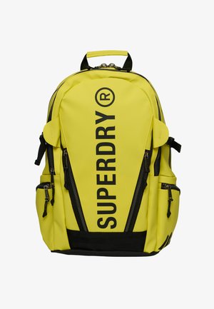 Felgele rugzak met zwarte verticale ritssluitingen en groot zwart "SUPERDRY" logo aan de voorkant, meerdere zijvakken met rits en een bovenhandvat.