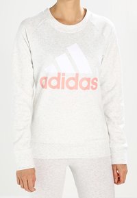 Persona che indossa un felpa Adidas grigia chiara con un grande logo bianco e rosa pallido sul petto.
