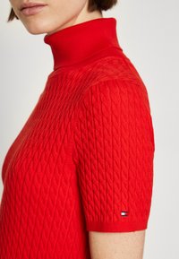 Pull rouge en tricot avec un col montant, présentant un design en câble texturé et des manches courtes. Comprend un petit logo sur la manche.