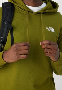Groene hoodie met een wit logo op de borst, voorzien van een trekkoord op de capuchon en geribbelde boorden. Draag met een zwarte rugzakriem zichtbaar.