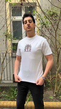 Νέος άνδρας που φοράει λευκό t-shirt Santa Cruz Skateboards και μαύρο παντελόνι στέκεται έξω κοντά σε γυμνά δέντρα και ένα παράθυρο κτιρίου.