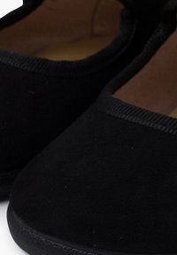 Zapatillas elásticas negras tipo slip-on con una textura suave similar al ante, puntera redondeada y un delgado ribete negro alrededor de la apertura.