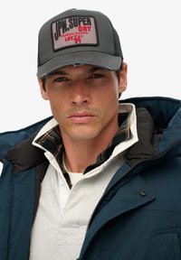 Hombre con gorra gris y roja, chaquetas en capas con abrigo azul marino y cuello de cuadros, mirando al frente con expresión neutral.