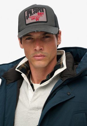 Hombre con gorra gris y roja, chaquetas en capas con abrigo azul marino y cuello de cuadros, mirando al frente con expresión neutral.