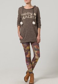 Maglione lavorato a maglia marrone con cordoncino e pompon, abbinato a leggings multicolore con pattern e stivaletti marroni alla caviglia.
