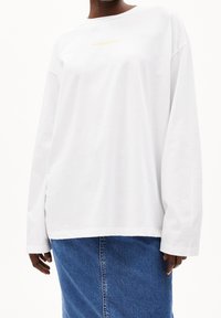 Persona che indossa una camicia bianca a maniche lunghe e ampia con testo giallo chiaro e una gonna di jeans blu, mostrata dalla gola fino ai fianchi.