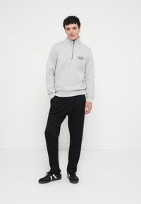 Grauer Kapuzenpullover mit Reißverschluss und hohem Kragen, mit einem kleinen schwarzen Logo auf der Brust, kombiniert mit lockeren schwarzen Hosen und schwarzen Turnschuhen.