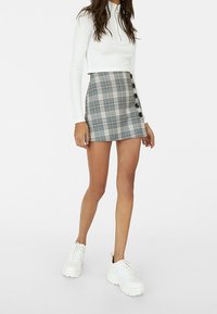 Femme portant un crop top blanc à manches longues, une mini-jupe grise à carreaux avec boutons et des baskets blanches épaisses, debout sur un fond blanc.