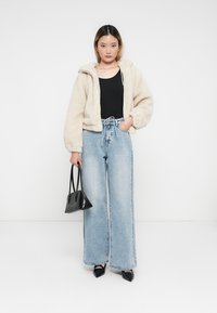 Γυναίκα με μπεζ φλις ζακέτα, μαύρο τοπ, φαρδιά μπλε τζιν παντελόνι, μαύρες γόβες, κρατώντας μαύρη τσάντα, στέκεται μπροστά σε λευκό φόντο.