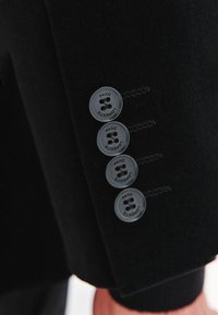 Zbliżenie na rękaw czarnego garnituru z czterema czarnymi guzikami oznaczonymi napisem "PAKO LORENTE", naszytymi w pionowej linii.