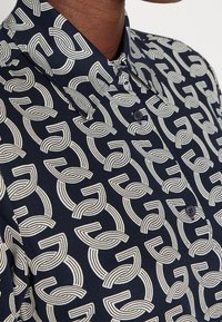 Gros plan d'une chemise bleu foncé à boutonnage avec un motif répétitif de lignes courbes blanches abstraites portée par une personne à la peau foncée.