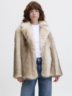 Young person with medium-length brown hair wearing a beige and cream faux fur coat, white shirt, and blue jeans against a plain background.