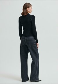 Svart ribbad tröja med åtsittande design, parat med högmidjade mörka jeans. Modellen står med ryggen vänd mot kameran mot en ljus bakgrund.