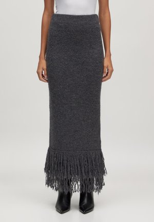 Donkergrijze gebreide lange rok met franjes aan de zoom, gedragen met zwarte puntige laarzen en een wit topje, afgebeeld van middel tot voeten.