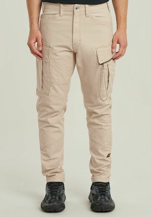 Beige Cargo-Hosen mit mehreren Taschen, einer seitlichen Tasche mit Reißverschluss und einem schmal zulaufenden Schnitt. Hergestellt aus haltbarem Stoff mit sichtbaren Nähten.