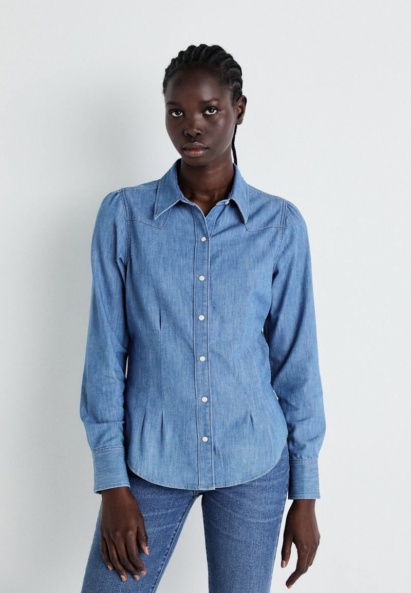 Jasnoniebieska koszula dżinsowa z wyłogowym kołnierzykiem, z przodem zapinanym na guziki oraz długimi rękawami. Posiada plisowane detale i zaokrąglony dół.