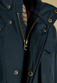 Detailný záber na čiastočne zipsovanú tmavomodrú bundu so mosadzným zipsom a cvokmi, ktorá je oblečená cez vzorovaný sveter.