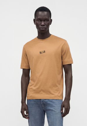 Homme à la peau foncée et aux cheveux courts porte un t-shirt marron à manches courtes avec un petit logo sur la poitrine et un jean bleu, debout devant un fond blanc.