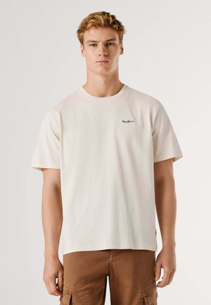 Jeune homme aux cheveux blonds courts portant un t-shirt crème uni et un pantalon cargo marron, debout devant un fond clair.