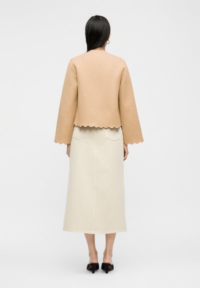 Γυναίκα με μακριά μαύρα μαλλιά φοράει μπεζ σακάκι με κυματιστό τελείωμα, κρεμ μίντι φούστα με πίσω τσέπες και μαύρες ψηλοτάκουνες μύτες, στέκεται γυρισμένη προς τα πίσω.