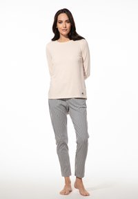 Vrouw die blootsvoets staat en een lichtbeige shirt met lange mouwen en blauw-witte verticaal gestreepte broek draagt, tegen een effen achtergrond.