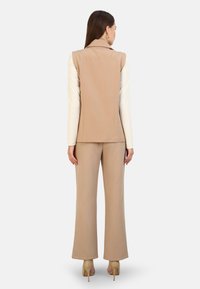Beige sako a odpovídající široké kalhoty vyrobené z hladkého materiálu; obsahuje límec a elegantní design. Klasická obuv doplňuje vzhled.