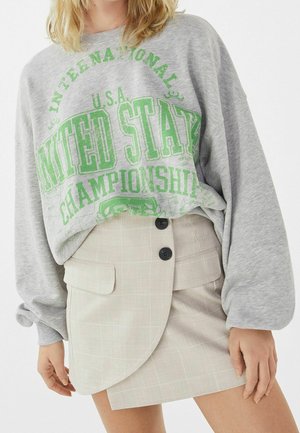 Person iført en stor grå sweatshirt med grøn "United States Championship" tekst og beige ternet asymmetrisk knapbundet nederdel.