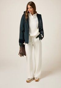 Giacca in shearling di un verde scuro oversized con cappuccio, felpa bianca e pantaloni jogger, guanti verde scuro, borsa marrone e sneakers beige e marroni.