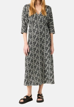 Mujer vestida con un vestido midi estampado en blanco y negro con mangas tres cuartos y sandalias negras con hebillas, de pie frente a un fondo liso.