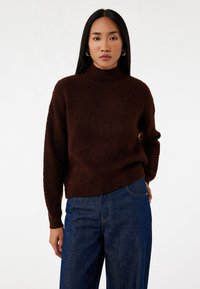 Brun strikket sweater med høj krave, præget af et tekstureret mønster og løst fit. Sammen med højtaljede denimjeans.