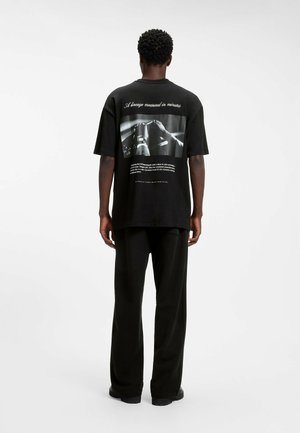Mann trägt schwarzes übergroßes T-Shirt mit schwarz-weißem Foto und Text auf dem Rücken, kombiniert mit lockeren schwarzen Hosen, steht vor weißem Hintergrund.