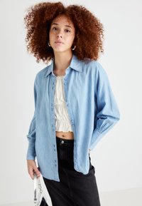 Lyseblå denimskjorte med knapfront, lange ærmer og kravehals, lagd over en kremfarget bluse med volanger. Svart skjørt og skulderveske.