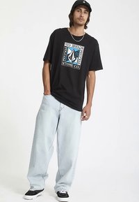 Zwart grafisch T-shirt met een kleurrijk logo-ontwerp, gecombineerd met lichtblauwe baggy jeans en zwart-witte sneakers. Casual streetwear stijl.