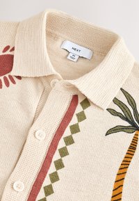 Beige strikket sweater med polo-krave. Har røde blomster- og grønne palmebladmotiver, fremhævet med mønstrede striber. Knappelukning.