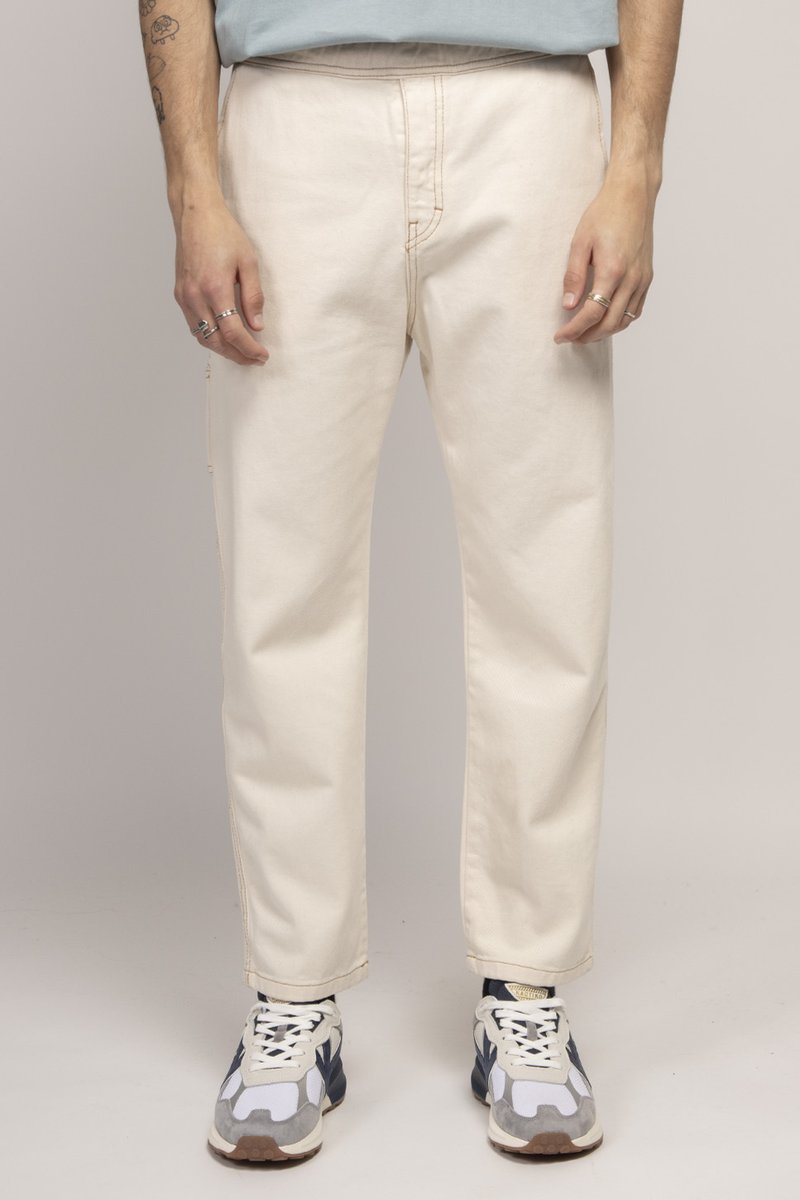 Pantalons en coton beige clair avec une coupe droite, des coutures visibles et une texture subtile. Associés à des baskets grises et blanches.