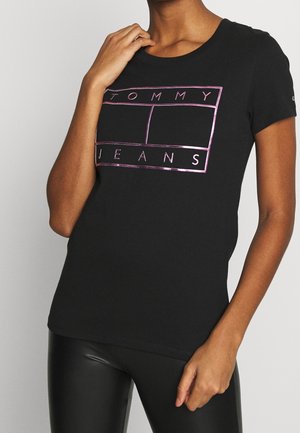 Henkilö, joka pukeutuu mustaan istuvaan T-paitaan, jossa on metallinhohtoinen pinkki "TOMMY JEANS" -teksti ja ääriviivat, yhdistettynä mustiin nahkalegginseihin.