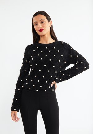 Donna con capelli scuri che indossa un maglione nero decorato con perle bianche distribuite uniformemente e pantaloni neri aderenti su sfondo bianco.