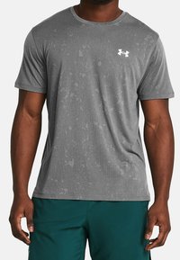 Šedé atletické tričko s krátkým rukávem vyrobené z texturované látky, se subtilním abstraktním vzorem a bílým logem na levé straně hrudi.