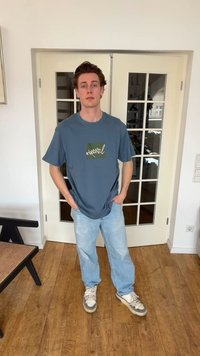 Νέος άνδρας στέκεται μέσα σε εσωτερικό χώρο με τα χέρια στις τσέπες, φορά μπλε μπλουζάκι με σχέδιο, ανοιχτόχρωμο μπλε τζιν και λευκά αθλητικά, μπροστά από γυάλινες πόρτες.