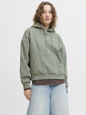 Ung person med krøllete hår iført en grønn hettegenser med frontlomme og lyseblå jeans, stående foran en enkel hvit bakgrunn.