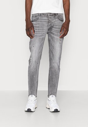 Mand iført slank grå distressed jeans med subtile malingsprøjt og hvide atletiksko mod en ensfarvet baggrund.