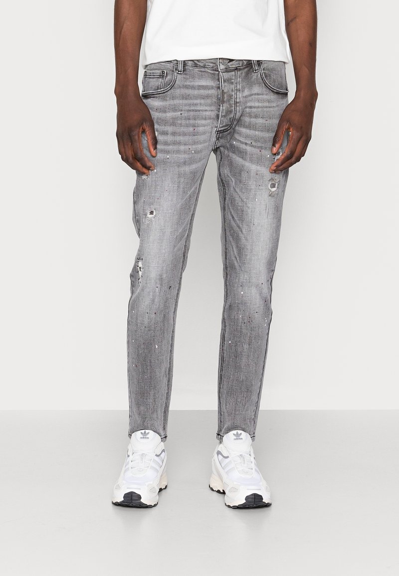 Homme portant un jean slim gris usé avec de subtiles éclaboussures de peinture et des baskets blanches sur un fond uni.